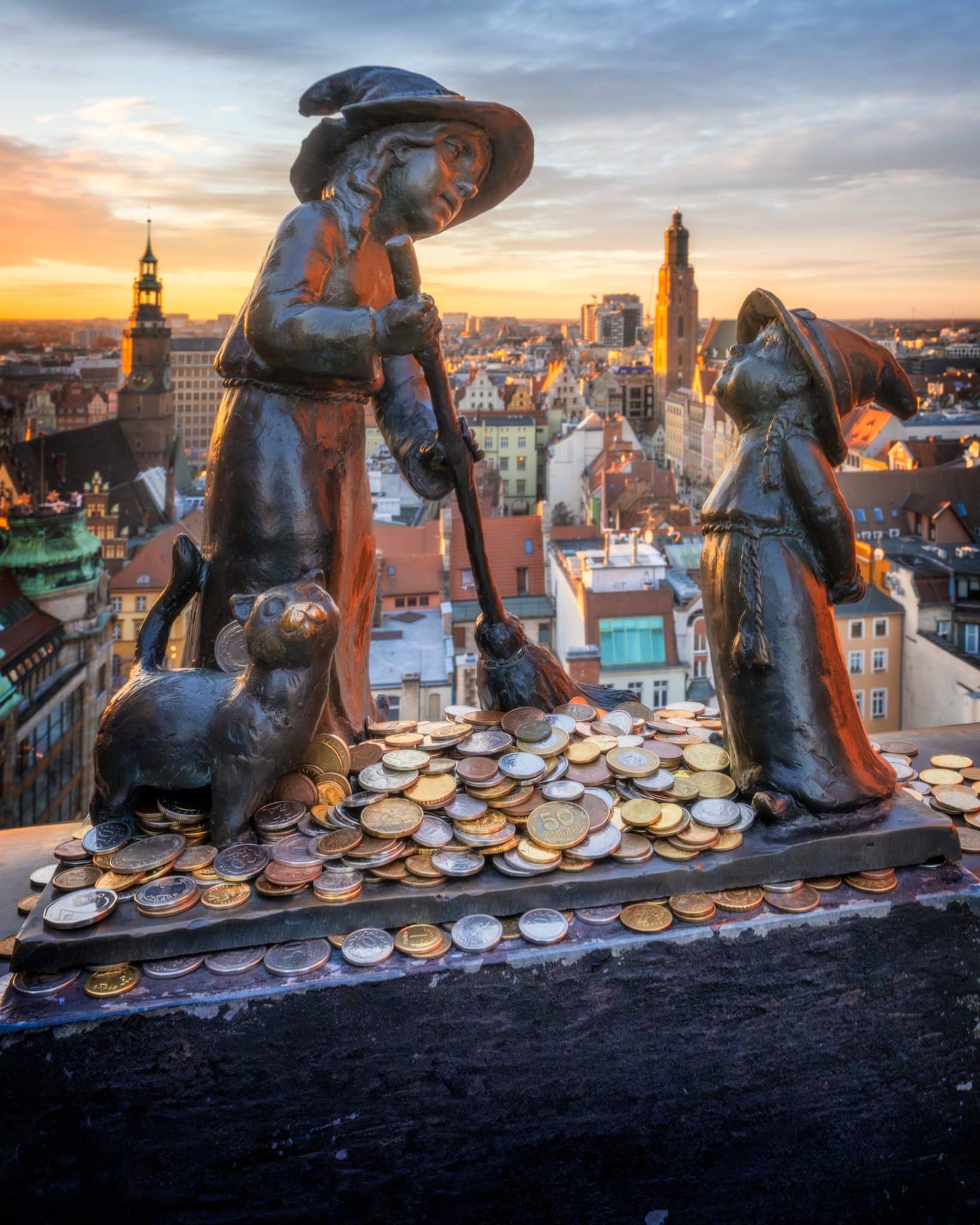 Bronze witch statues and a cat surrounded by coins on Wroclaw's Witches' Bridge at sunset.