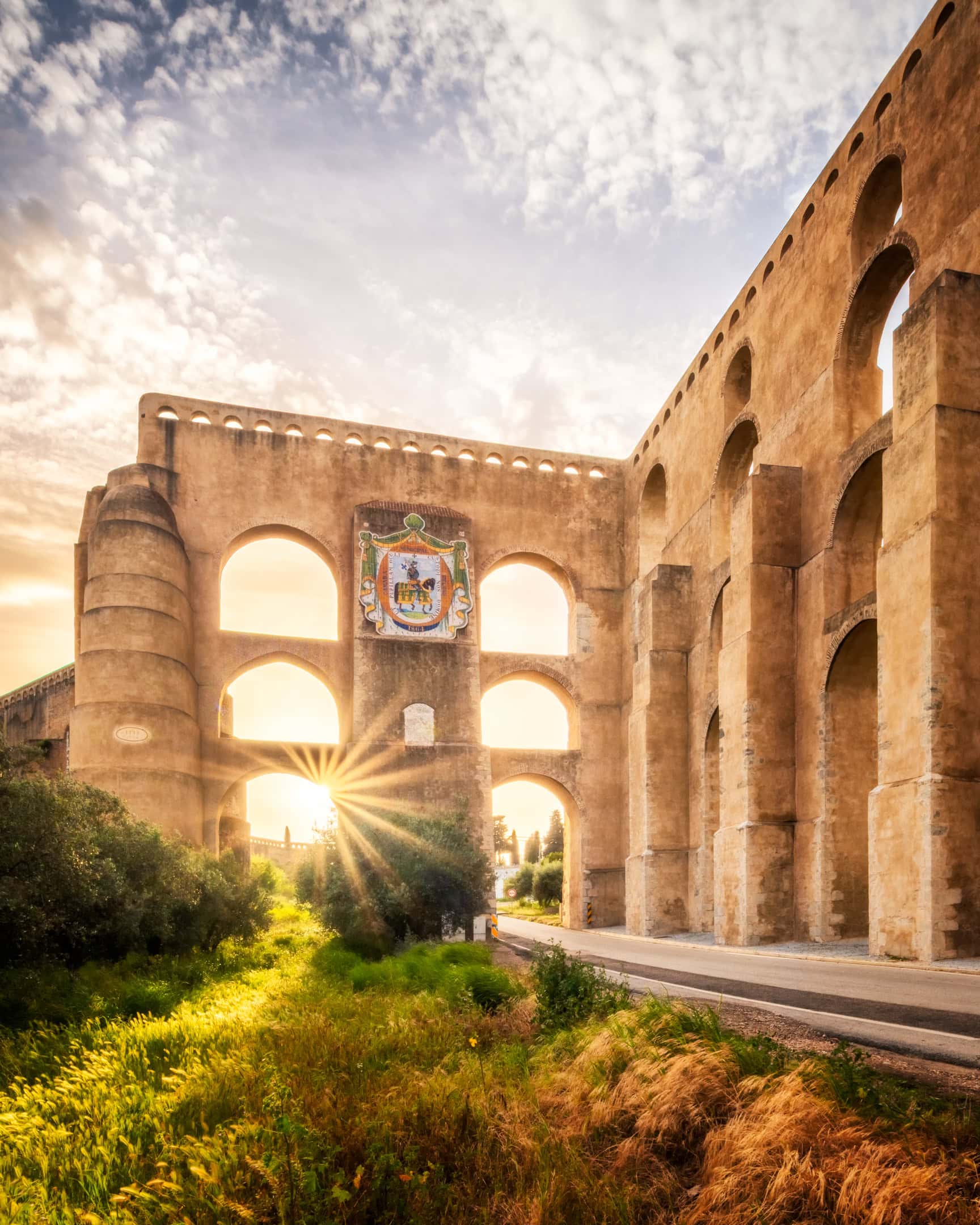 Amoreira Aqueduct in Elvas Portugal, sunlight bursting beneath its tallest arch tower crowned by a painted coat of arms