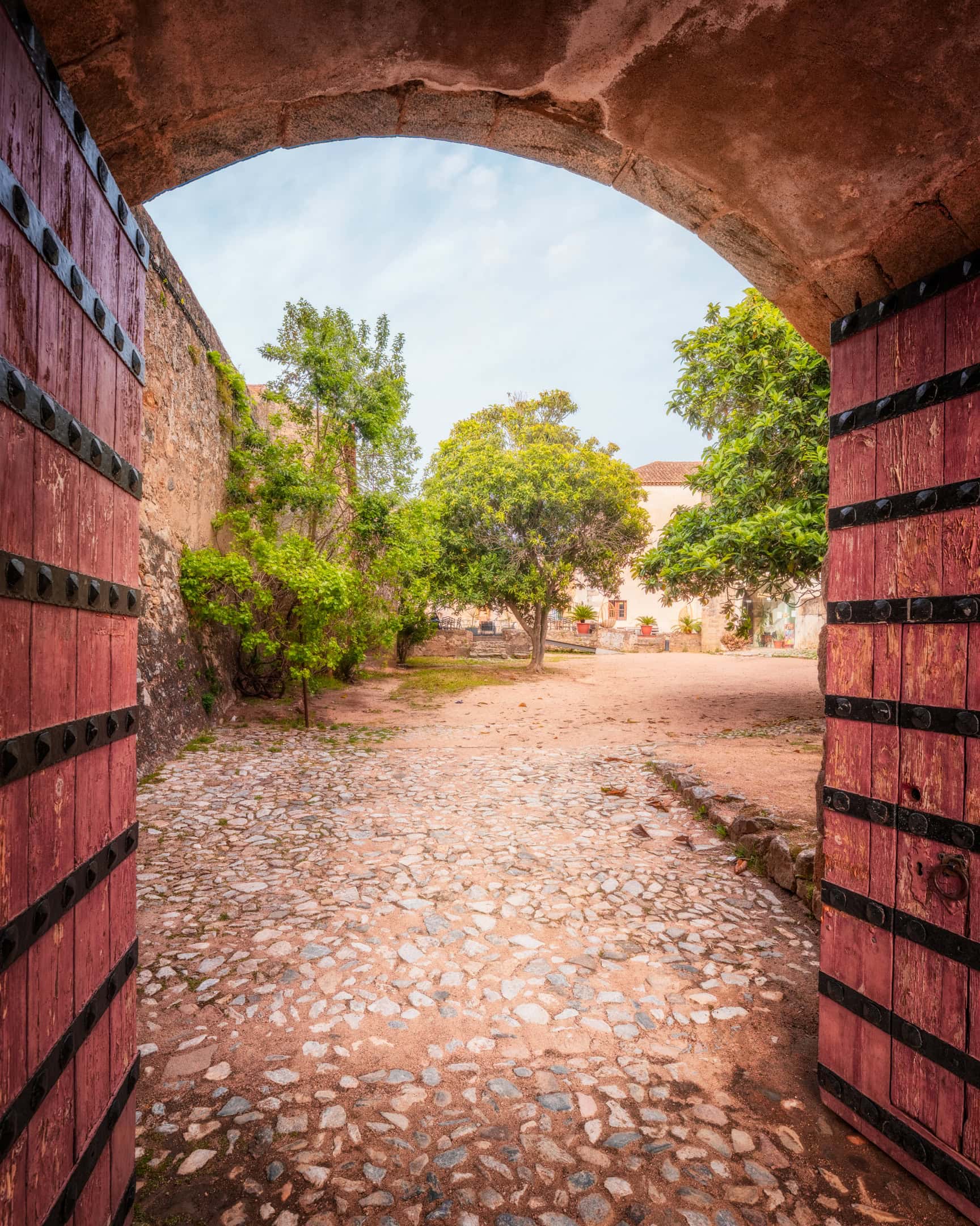 View through the studded red wooden gate of Elvas Castle into the tree shaded inner courtyard, Elvas, Portugal